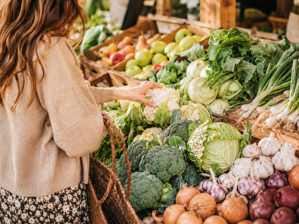 Eine Frau mit einer Einkaufstasche am Arm steht vor einem Marktstand mit Gemüse und blähenden Lebensmitteln wie Brokkoli, Kohl, Zwiebeln und Knoblauch und greift einen Blumenkohl um ihn zu kaufen.