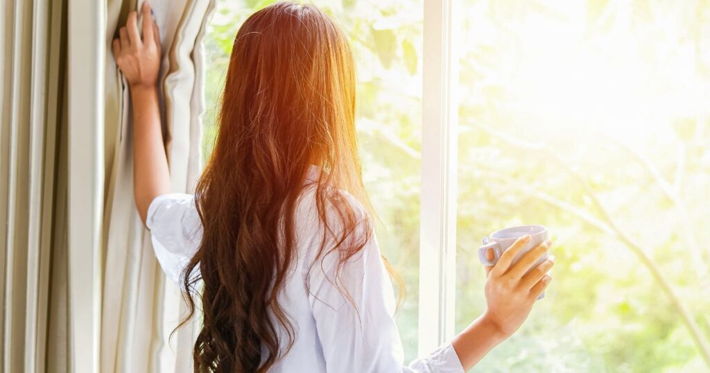 Frau steht mit einer Tasse Tee am Fenster, hält den Vorhang zur Seite und lässt Sonnenlicht herein.
